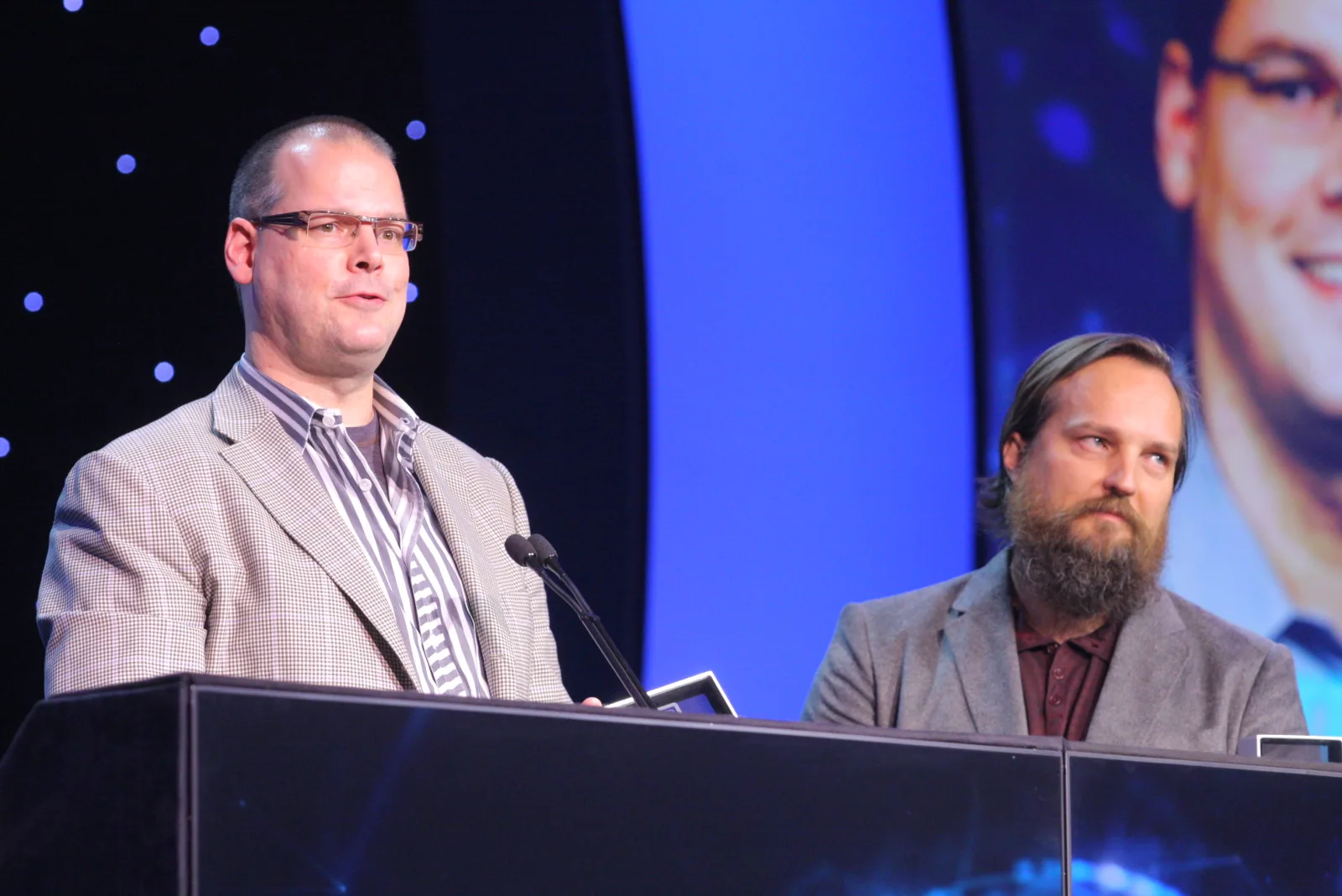 Die BioWare-Gründer Ray Muzyka (links, kariertes Sakko) und Greg Zeschuk (rechts, Vollbart) stehen an einem Rednerpult auf einer blau beleuchteten Bühne. Muzyka hält eine Auszeichnung in der Hand.